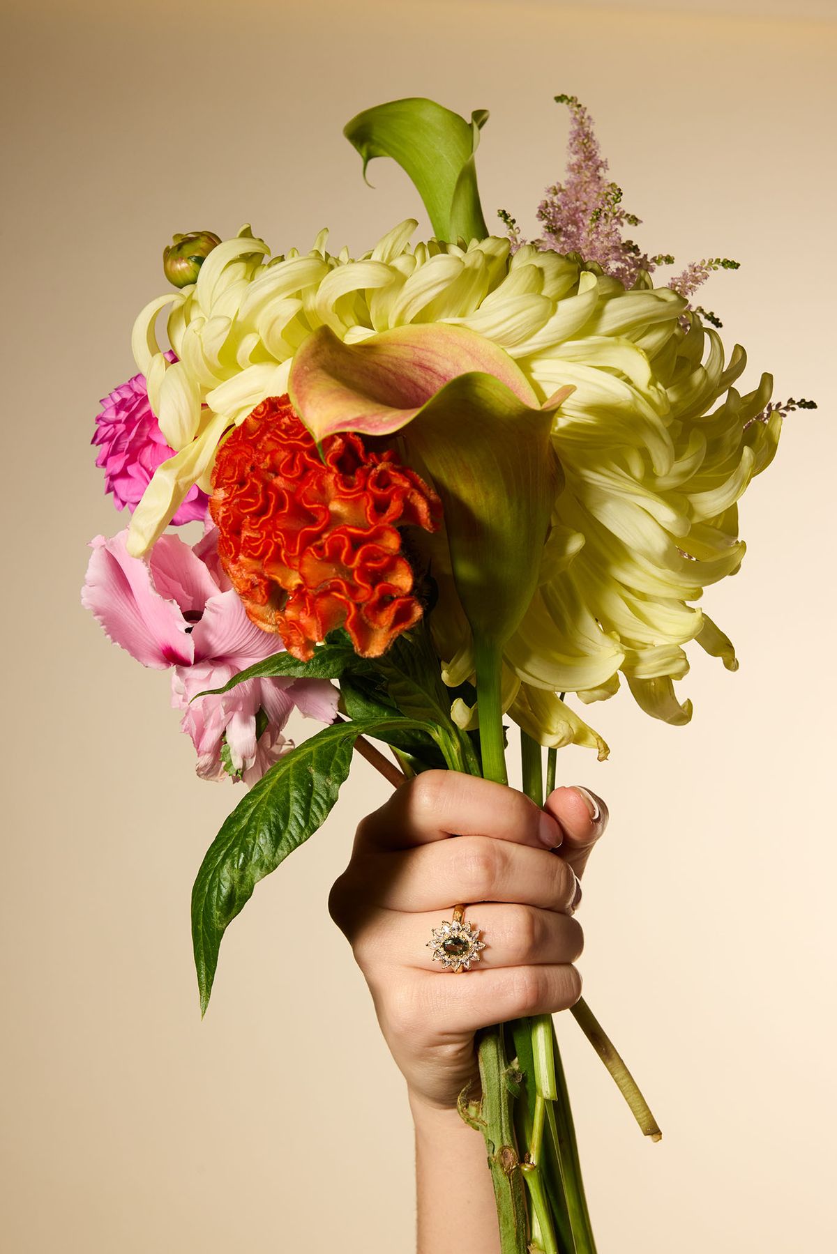 Une main féminine présente un bouquet vibrant de fleurs, comprenant des callas, des chrysanthèmes et d'autres variétés, dans un fond clair. La scène évoque une atmosphère joyeuse.